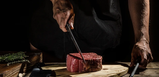 A close-up of a chef's hands slicing into a thick raw steak on a wooden board, set against a dark, moody backdrop with herbs nearby.