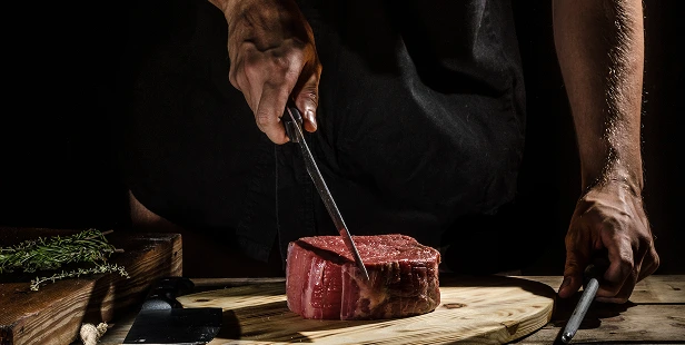 A chef slices through a thick cut of raw steak on a wooden cutting board, with herbs and tools visible in the moody, dark-lit setting.