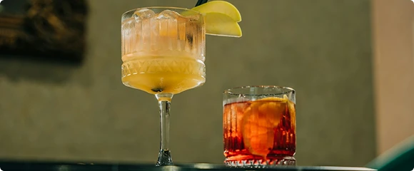Two craft cocktails sit side by side — a tall stemmed glass with a citrus garnish and a short rocks glass with an amber drink.