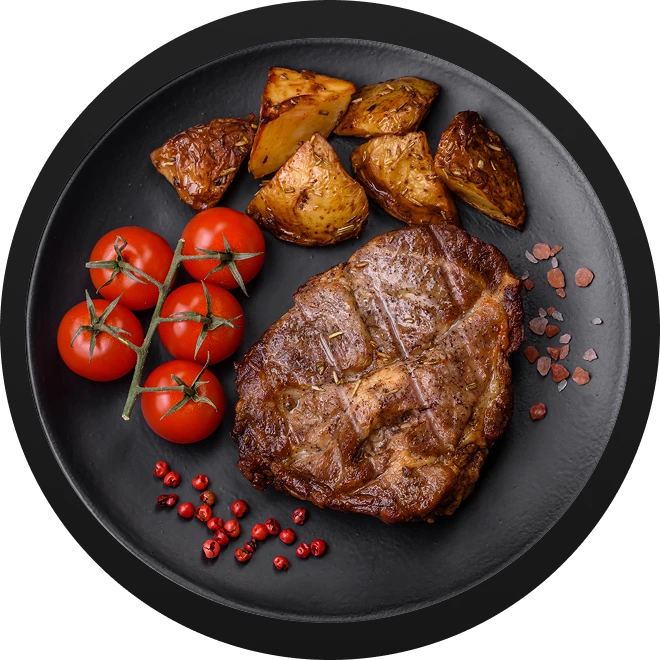 A perfectly grilled steak served on a dark plate alongside cherry tomatoes on the vine and roasted potatoes, with scattered pink peppercorns.