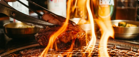 A piece of meat is held with tongs over a round charcoal grill, with bright orange flames rising around it.