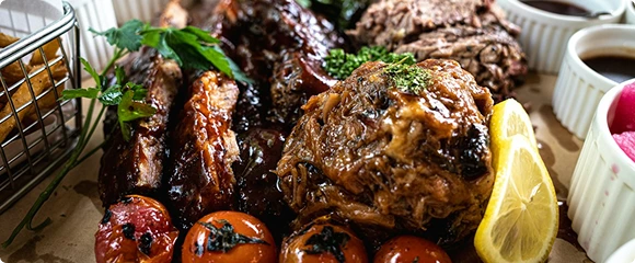 A generous platter of slow-roasted meat garnished with herbs, roasted tomatoes, and lemon, served alongside small side dishes.