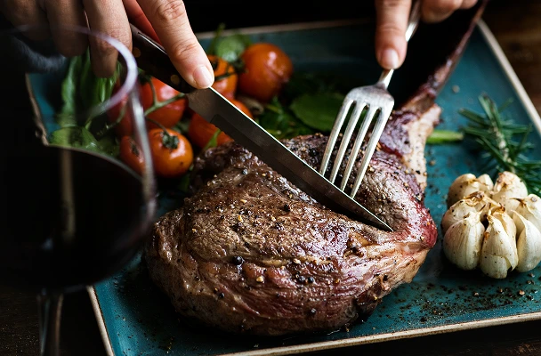 A chef uses a spatula and tongs to flip a seared steak on a commercial flat-top grill, captured in warm, close-up detail.