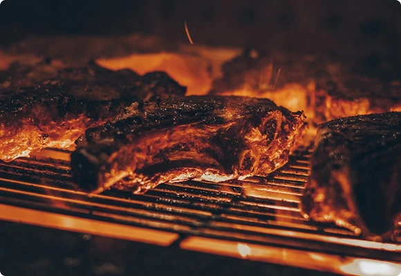 Racks of ribs cook slowly on a grill grate, with glowing embers and flames visible beneath the darkened, smoky meat.