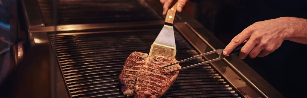 A wide shot of a steak being flipped with a spatula on a large commercial grill, with the chef's hands and tools in frame.